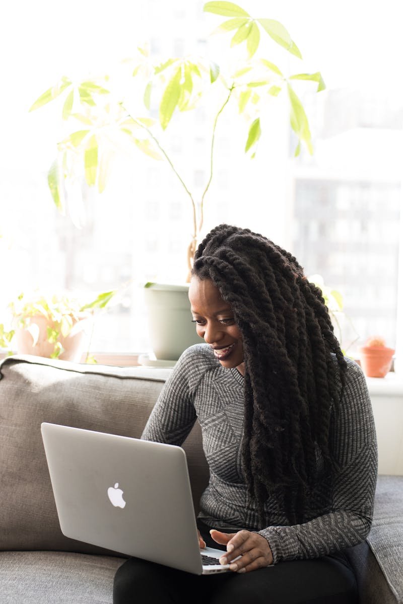 Black woman sitting on a sofa, smiling while working on a laptop indoors.